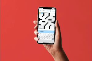 A brown hand holding a smartphone with The New York Times daily crossword loaded on it and visible on the screen, against a cherry red background.